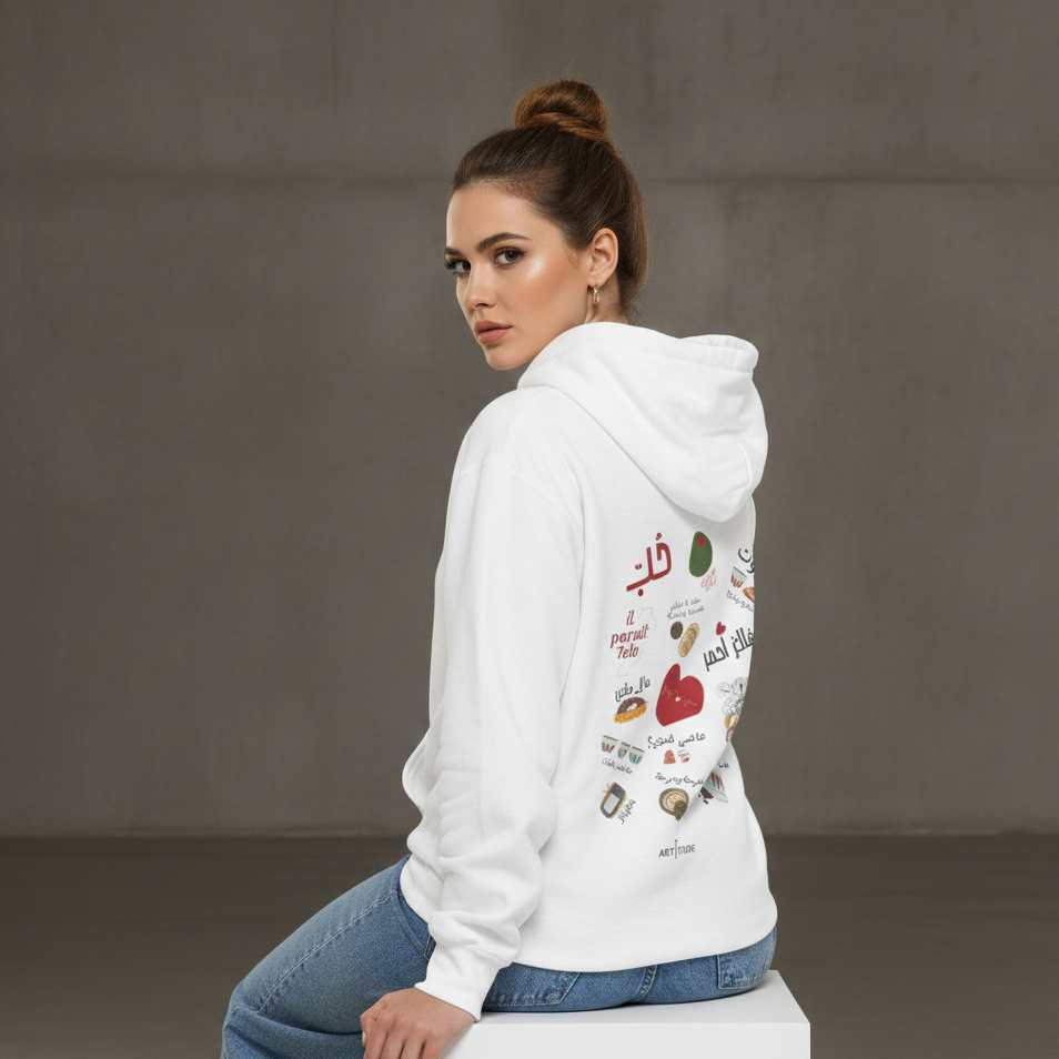 Woman wearing a white hoodie with Arabic text and graphics, sitting on a white box against a gray background.