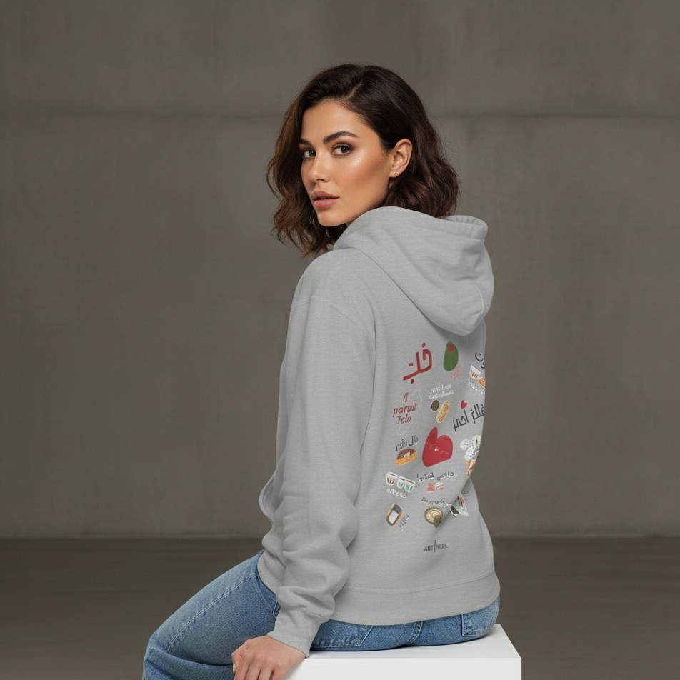 Woman wearing a gray hoodie with Arabic text and graphics, sitting on a white box against a plain background.