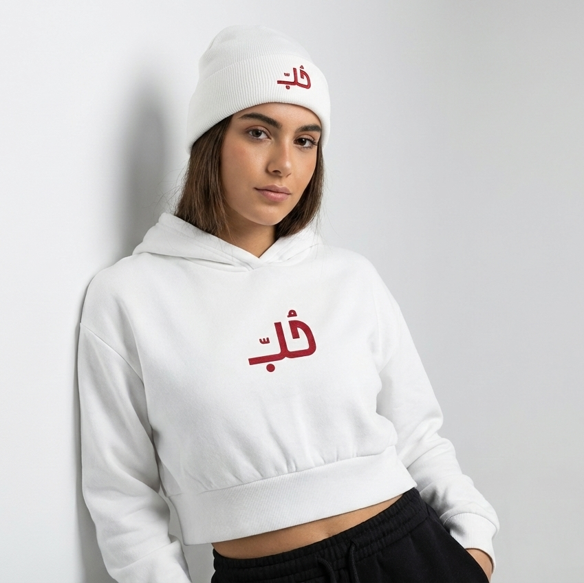 Person wearing a white hoodie and cap with Arabic text on a plain background