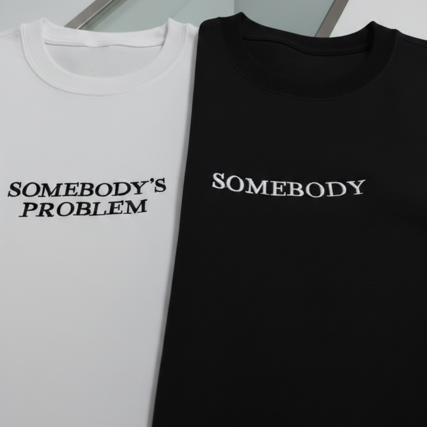 Two shirts, one white and one black, with text 'SOMEBODY'S PROBLEM' on a neutral background.