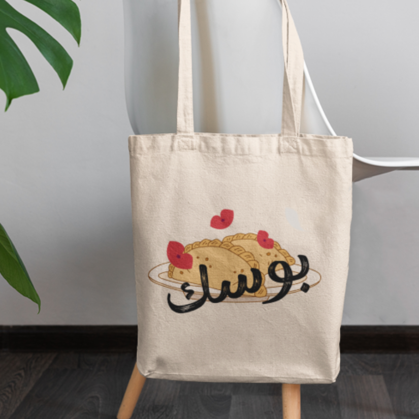 Tote bag with a design of a pastry and Arabic text on a wooden stand, with a plant in the background.