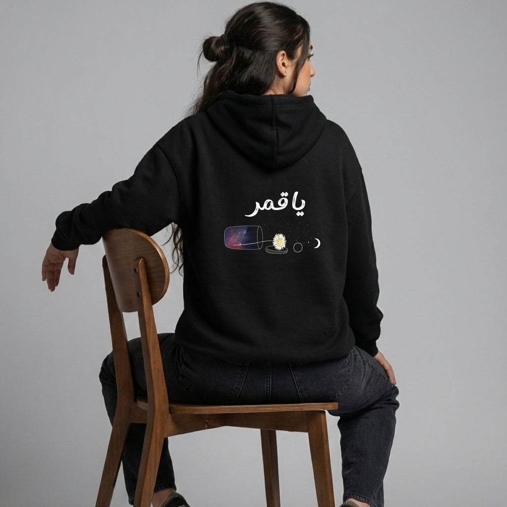 Person wearing a black hoodie with Arabic text and graphics, sitting on a wooden chair against a gray background.